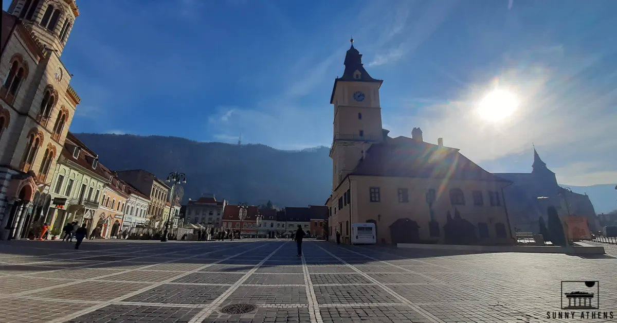 Unique Experiences in Brasov: The Council Square is one of the must stops of your 2 days in Brasov itinerary. In the picture, two people are walking the stone paved square under the sunlight.