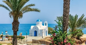 Best day trips from Athens: a traditional white and blue Greek chapel by the sea, framed by tall palm trees and vibrant pink flowers.