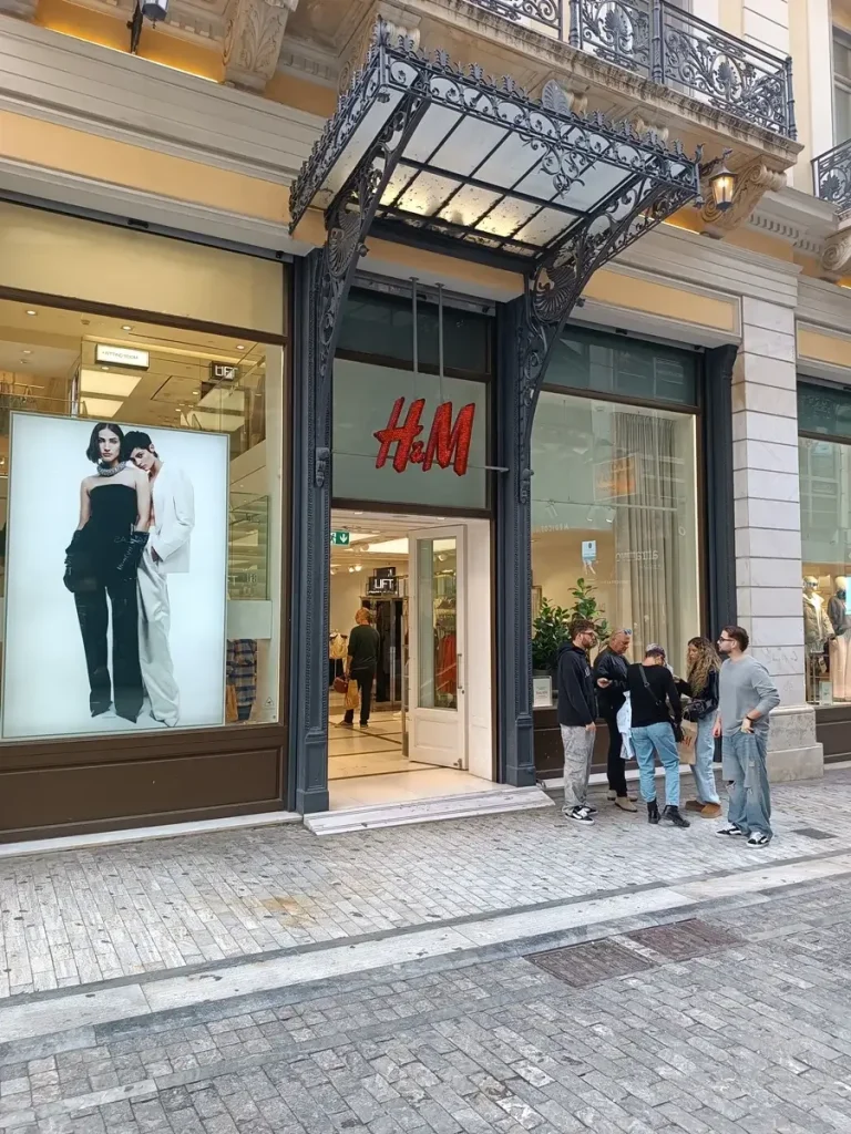 The entrance of H&M store at Ermou Street in Athens, with people standing outside and discussing.