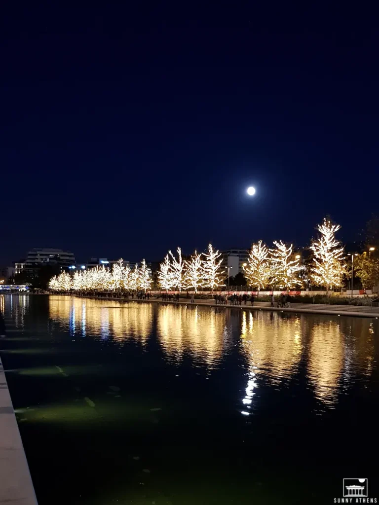 Nighttime view at the Stavros Niarchos Foundation Cultural Center, featuring illuminated trees reflecting on the water, and a full moon shining brightly.