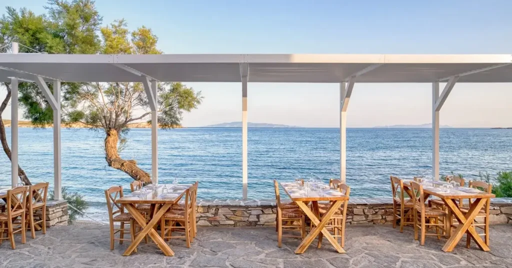 Wooden tables of the gourmet Kima restaurant, under a white kiosk and with a green tree in the left background, by the sea in Drios, Paros.