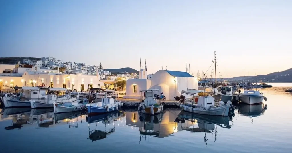 The picturesque port of Naousa in Paros, Greece, featuring a tiny white church and small fishing boats at dusk.