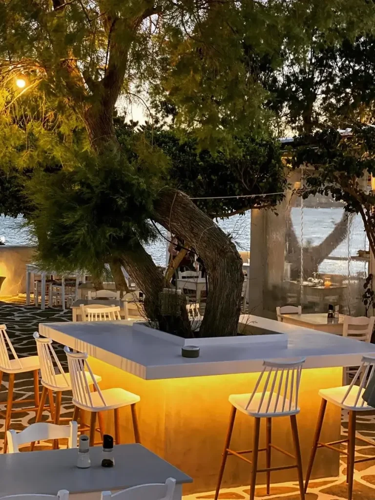 Outdoor seating at Siparos Seaside Restaurant in Paros, with white tables and chairs under a green tree, overlooking the sea at dusk.