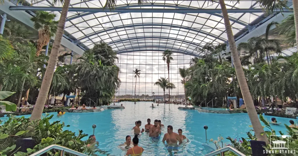 People swimming in the main pool of Therme Bucharest, under a vast glass roof with trees surrounding them.