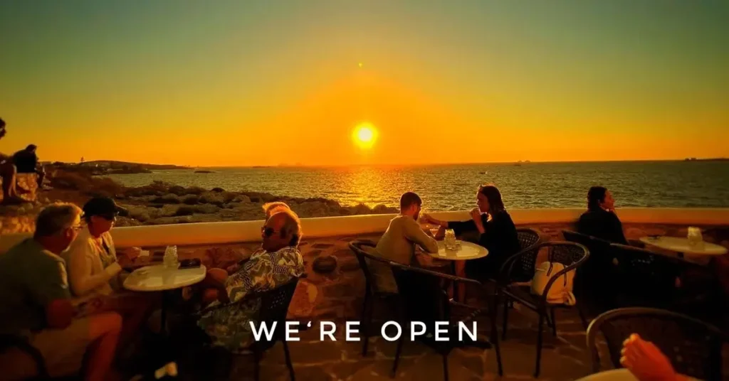 People seated in the yard of Alexandros Cafe Bar in Paros, Greece, at dusk, enjoying the sunset and a unique view of the Aegean Sea.