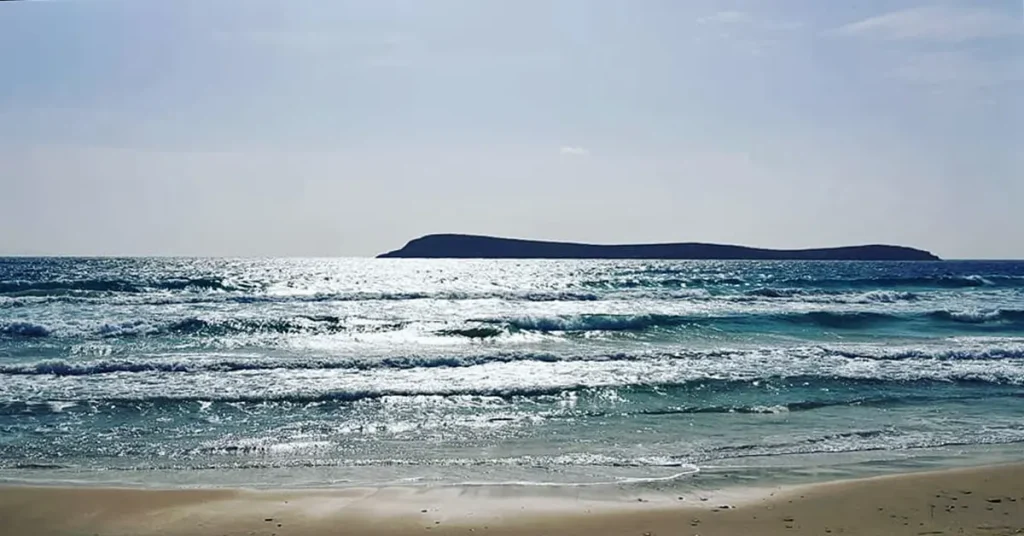 Small waves breaking on the sandy Parasporos Beach with a view of a distant island under the summer Greek sun.