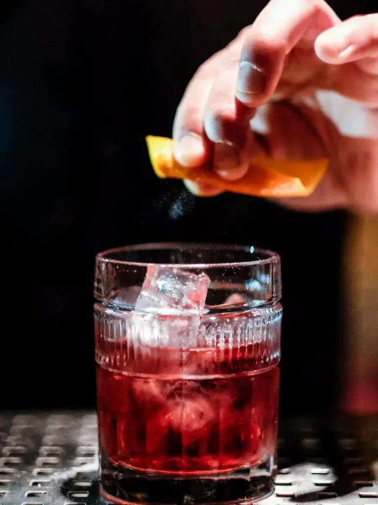 A hand squeezing an orange peel over a glass of Negroni, infusing the cocktail with a hint of orange essence.