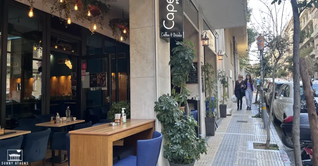 A café with outdoor seating and plants on Skoufa street in Kolonaki, Athens, with two women walking by.