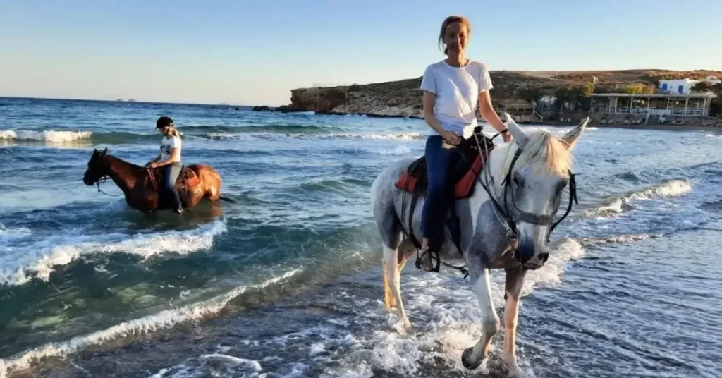 Two women riding a white and a brown horse by the sea in Paros Greece.