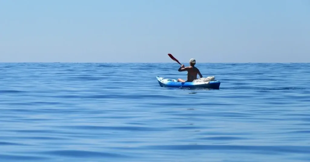 A man paddling a blue kayak alone in the calm blue sea under clear skies.